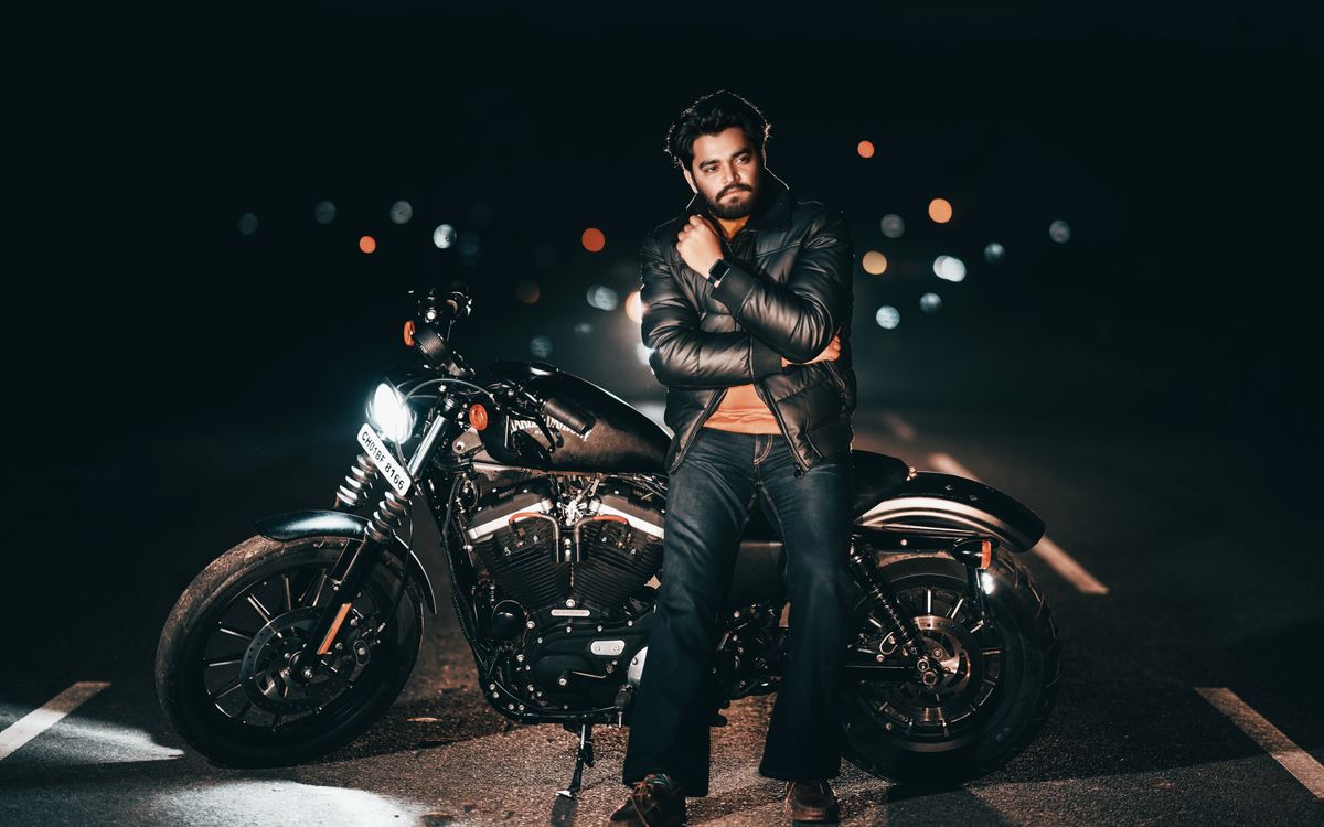 Buneri.es | Bomber, motera o café racer: ¿qué estilo de chaqueta de cuero te queda mejor? Hombre con chaqueta de cuero posando junto a una moto de noche con luces de la ciudad de fondo