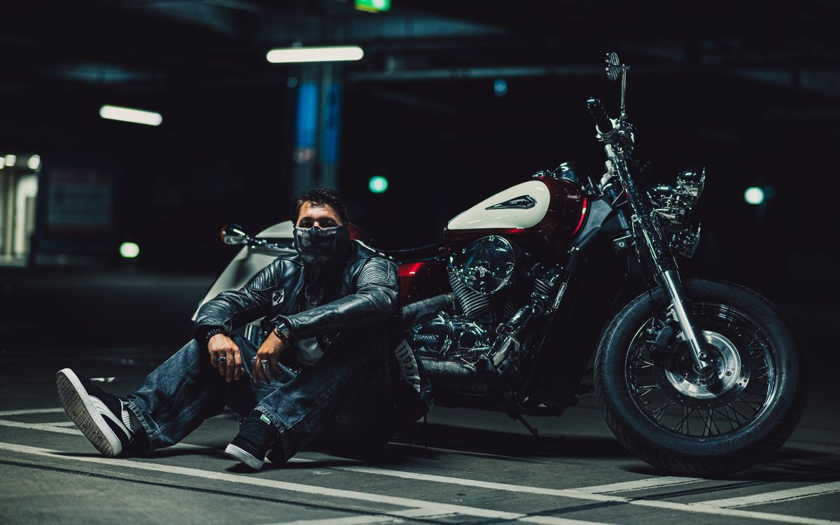 Motero en chaqueta de cuero arrodillado junto a una moto en un garaje poco iluminado